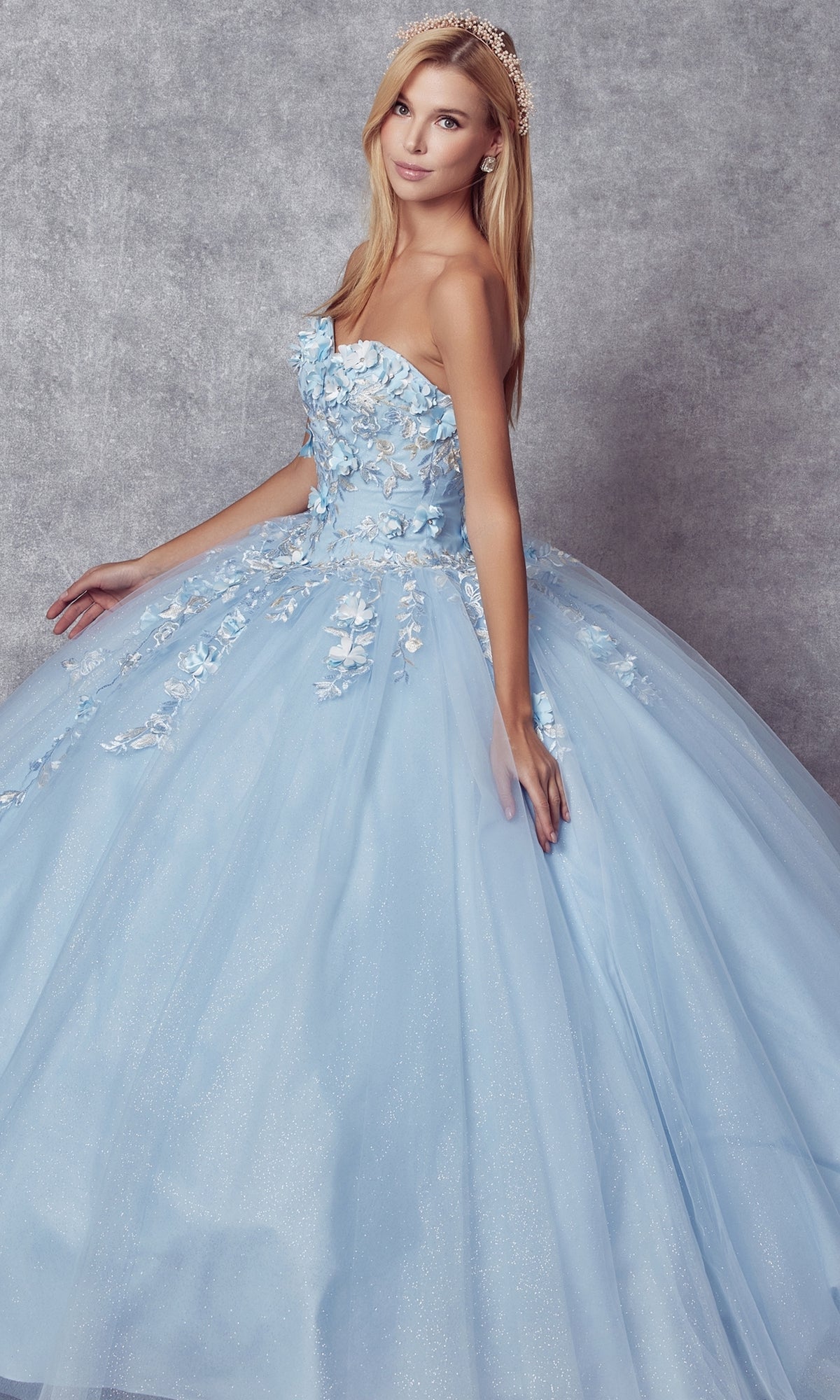 A woman models the Juliet 1435 Strapless Ball Gown Quinceañera Dress in light blue with floral embellishments, standing against a gray textured background and capturing its elegant style.